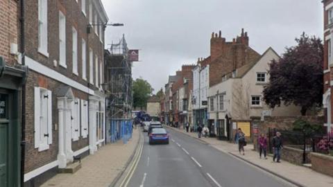 A road with buildings on either side. Scaffolding covers the front of one building. People are walking on the pavement. A tree between two buildings can be seen.