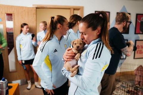 A woman in the foreground of the picture has her hair styled in a high ponytail and is dressed in light blue sports gear. She is holding a six-week old puppy and looking down at it. In the background there are four women who are also dressed in sporting clothes and a man in a black tshirt, glasses with tattoos on his arm appears to be holding a camera and looking to the right of the frame.