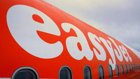 A picture of the side of an easyjet plane. It is orange and white in colour with the easyjet logo on the side of it.