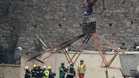 A crane lift rubble from an explosions site as emergency services in PPE look on.