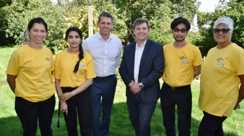 People involved in the project stand in a line as they are photographed outside. Two woman in yellow T-shirts stand next to a man in a white shirt.  Nik Jonson is in the middle and is wearing a blue suit and two more people in yellow T-shirts stand beside him. They are all smiling at the camera. 