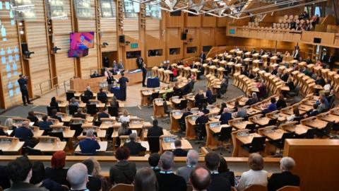 A wide shot of the Scottish Parliament chamber, with MSPs sitting at wooden desks in a wood-panelled room 