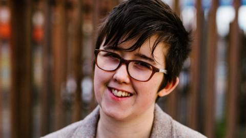 Lyra McKee, a woman with short dark hair, smiling at the camera with her head slightly tilted to one side. She is wearing brown-rimmed glasses and a beige tweed blazer. She is standing in front of a tall steel security fence. 