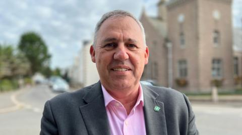 Constable Andy Jehan is a white man with short grey hair. He is wearing a pink shirt with a grey suit jacket and a pin of the right lapel. He is smiling into the camera. The background is an out-of-focus road and a granite parish hall.