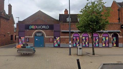 Exterior of a brick building with a black sign and multi-coloured writing with 'Popworld' on it. There is a tree, lamp post, bench and bin on the pavement