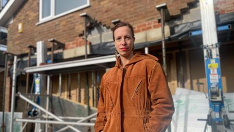 A woman in an brown suede coat stands with her hands in her pockets in front of a house covered in scaffolding.