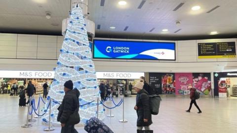 A large decorated and lit white Christmas tree inside a terminal at Gatwick Airport with warmly dressed travellers walking beside it