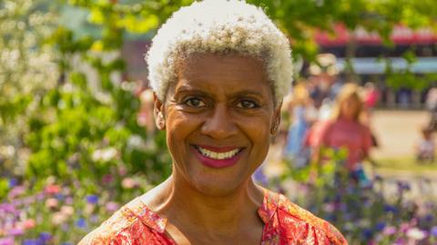 Arit Anderson is smiling and has short bleach blonde hair. She is wearing a colorful dress, and is standing in front of a garden.