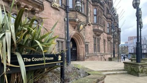 The council offices in Coventry are in a Tudor Revival-style building, built between 1913 and 1917, with an ornate exterior. A sign points to The Council House. People are walking around the building.