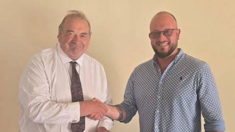 Two men are shaking hands - one is wearing a blue dotted short, and the other is wearing a white shirt with a red and blue tie.