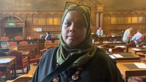 A woman is wearing a green hijab with a pair of glasses resting on top of her head. She has a black coat with a necklace and is standing in a wood panelled council chamber with wooden desks and chairs behind her