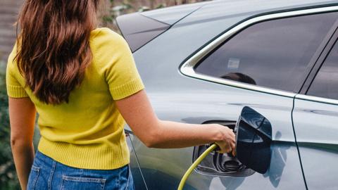 A young woman, wearing a bright yellow top is charging her electric car, parked in front of a house.