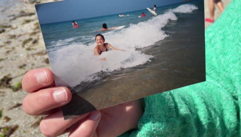 The picture is a close-up of a hand holding a photograph of a child on a surfboard in the sea. The person holding the old photograph is standing on a beach and is wearing a green jumper.