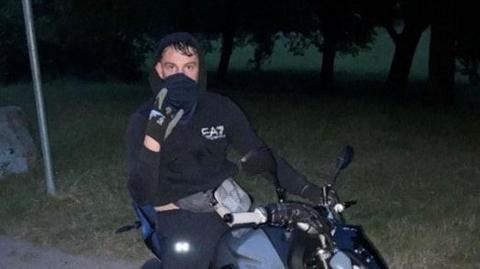 Kyle Camp, wearing a black hoodie and face covering, sits on a stolen motorbike. He is in a park and it is dark.