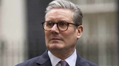 Headshot of Sir Keir Starmer. He stands outside what appears to be an out-of-focus Downing Street. He wears a dark jacket, a white shirt and a burgundy tie, with brown-rimmed spectacles and grey hair styled in a slight quiff.