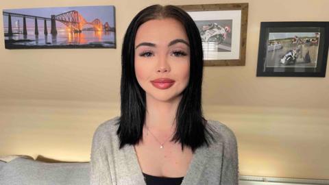 A woman with dark hair. She is wearing a grey cardigan.