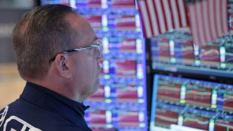 Trader wearing glasses looking to the right of picture, his gaze on multiple screens with USA flags above