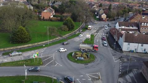 A roundabout in Bridlington, with lots of roads stemming from it. A lorry with a red cabin at the front is going round and there is a white building on one corner. The roundabout has a green grass island in the middle.