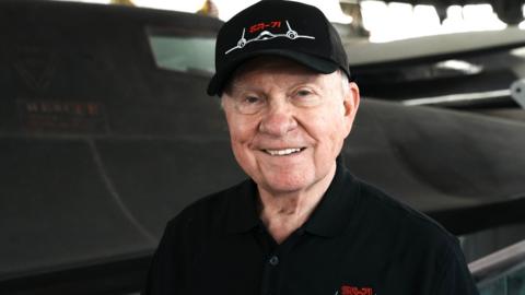 The head and shoulders of Buz Carpenter, standing in front of a Blackbird spy plane. He is wearing a black baseball cap and a black T-shirt with blackbird stitched on the left side. He is smiling broadly.