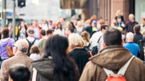 A sea of people to represent population. There are two people in the main frame, walking away from the camera. One has long dark hair and is wearing a green coat with a hood. The other has short brown hair and is wearing a brown coat with a hood and an orange backpack with white straps.