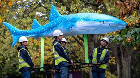 Four men in hard hats and fluorescent jackets carry a lantern shaped like a blue whale above them. Trees can be seen in the background with green and orange leaves 
