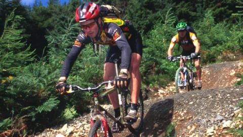 Two men on mountain bikes tackle undulating trails near Peebles on a tree-lined route.