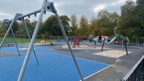 A large playground with black and blue flooring and surrounded by temporary fencing. An open van is parked nearby. 