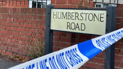 Blue and white police tape which says POLICE LINE DO NOT CROSS is across Humberstone Road in Luton. Within the police tape is a black and white road sign which reads HUMBERSTONE ROAD.