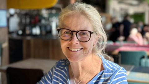 Jenny wearing a blue and white striped top. She is smiling and wearing spectacles. She is sitting inside.