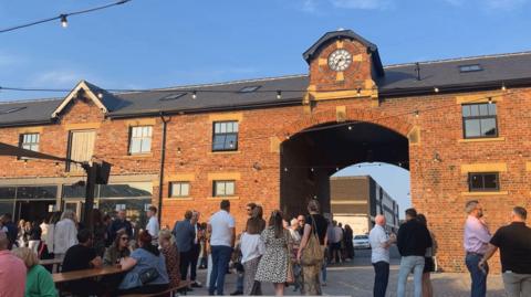 Dozens of people milling around on a sunny day outside a brick building