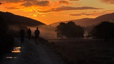 The silhouette of three men walking in front of a striking orange sky at sunrise. The orange tones are broke up with clouds. The men appear to be walking on a forrestry path, with puddles on it- which the is reflecting into. There are large black trees (also silhouetted) and mist in front of them.