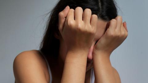 A teenage girl with olive skin and dark hair covers her eyes with her face in a crying pose.