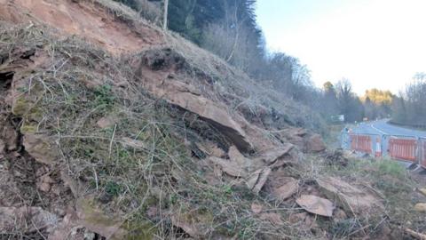 A slope of rocks near to a main road. The area has been cordoned off by gates and orange cones