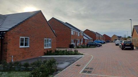 A picture of the housing development. There is a wide, paved road to the left of the picture, with a row of cars parked on it. On the right, there is a bungalow and a row of new houses. They are built from red brick and have white windows. Some have cars parked in front of them.