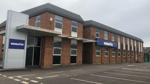 The exterior of the old Meco factory in Worcester. It is a brown brick building with a flat grey roof. Signs on the front and next to the building read "Komatsu". An empty car park is in front of the building.