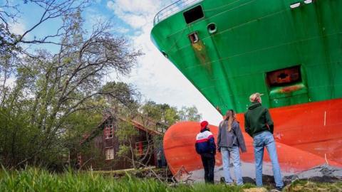 Three people look up at the green and red bow of a large container ship as it towers over a small wooden house that it narrowly avoided hitting