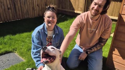 A photo of Charlotte and her partner Ryan kneeling down on grass behind Sasha the white Staffordshire bull terrier, which has its mouth open and its head arched to the sky. Everyone is happy. 