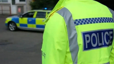Image of the back of a police officer wearing hi-vis jacket with a police car nearby outside a property. 