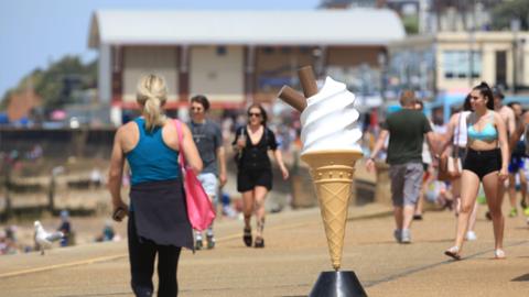 We can see people walking along a busy promenade in Hunstanton. In the foreground is a person-sized replica ice cream cone - in the background we can see a large pier amusements building.