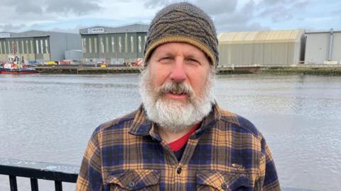 Dr Simon Gibbon is standing by the River Tees with industrial buildings on the far bank behind him. He's next to a black railing and is wearing a grey and mustard beanie hat. He has a short but luxuriant white beard, sideburns and moustache and is wearing a blue and mustard checked shirt with a red and black t-shirt underneath, just visible at the neck.