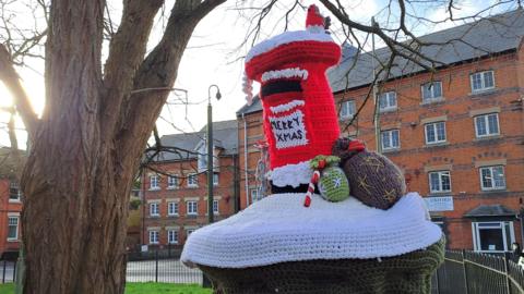 A knitted scene sits on top of a postbox. It includes a red postbox with a little robin on top. Next to it are two postbags and a candy cane. 