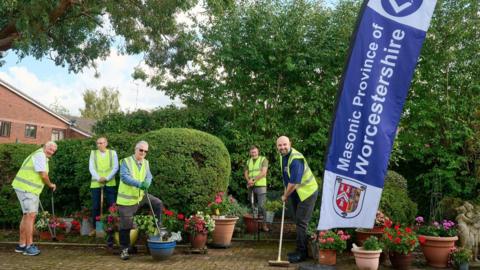 A group of five men in yellow high viz jackets carry out work in a garden area. The men pose next to a purple banner that reads: "Masonic Province of Worcestershire."