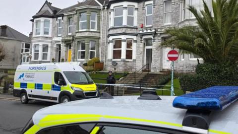 A police officer in uniform stands guard at a cordon outside a row of grey-bricked terraced houses in Plymouth. He is standing behind a line of police tape. A police forensics van is parked next to him on the road. The roof of a police car can also be seen in the foreground.