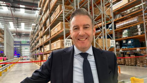 A man in a navy suit smiling in front of warehouse boxes all in columns.