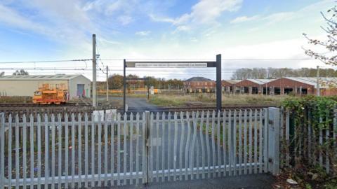 The Gosforth Metro depot in Newcastle. A metal gate bars access to the train yard. A number of its bars are bent. A sign warns people of high voltage overhead wires. A number of brick buildings can be seen in the yard which houses Metro trains.