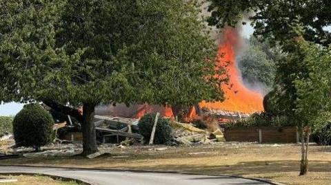 Fire at property in Devon. Trees in the foreground and a big blaze in the background with debris on the floor. Short grass and a drive in the foreground.