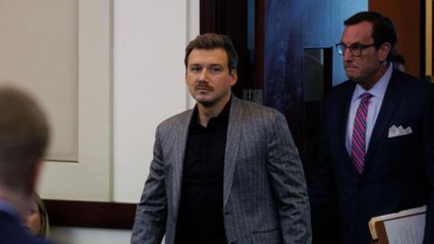 Man with moustache walks into room while wearing grey suit and black dress shirt