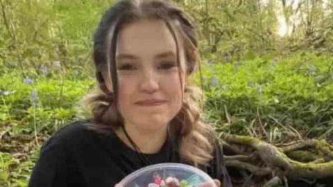 A teenage girl with brown hair smiling at the camera. She is wearing a black shirt, holding a bowl of sweets and is in a forest area.