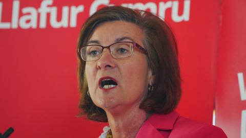 Eluned Morgan wearing a red jacket in front of a red Welsh Labour banner, with Llafur Cymru written in Welsh behind her