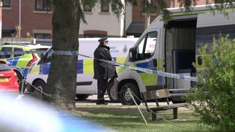 A crime scene at a park. Police tape has been attached to a tree and stretches out of frame. A police officer can be seen standing on the side of a road next to several police vehicles.
